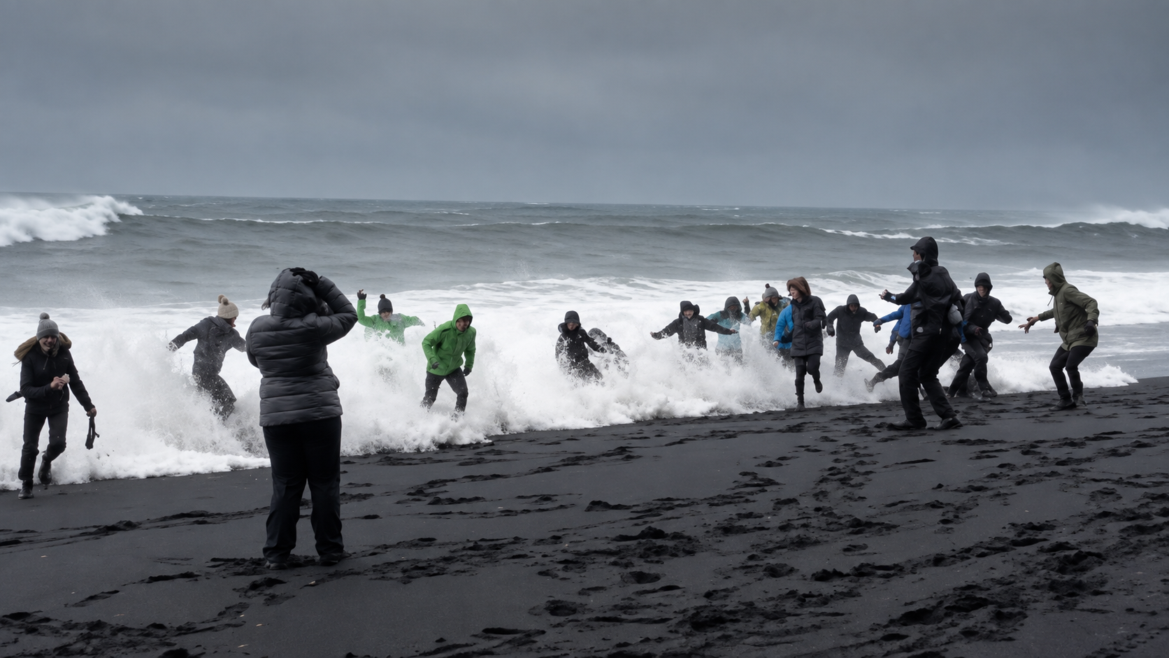 A group of people in jackets run from incoming ocean waves on a black sand beach under cloudy skies, with one person standing closer to the camera capturing the scene.