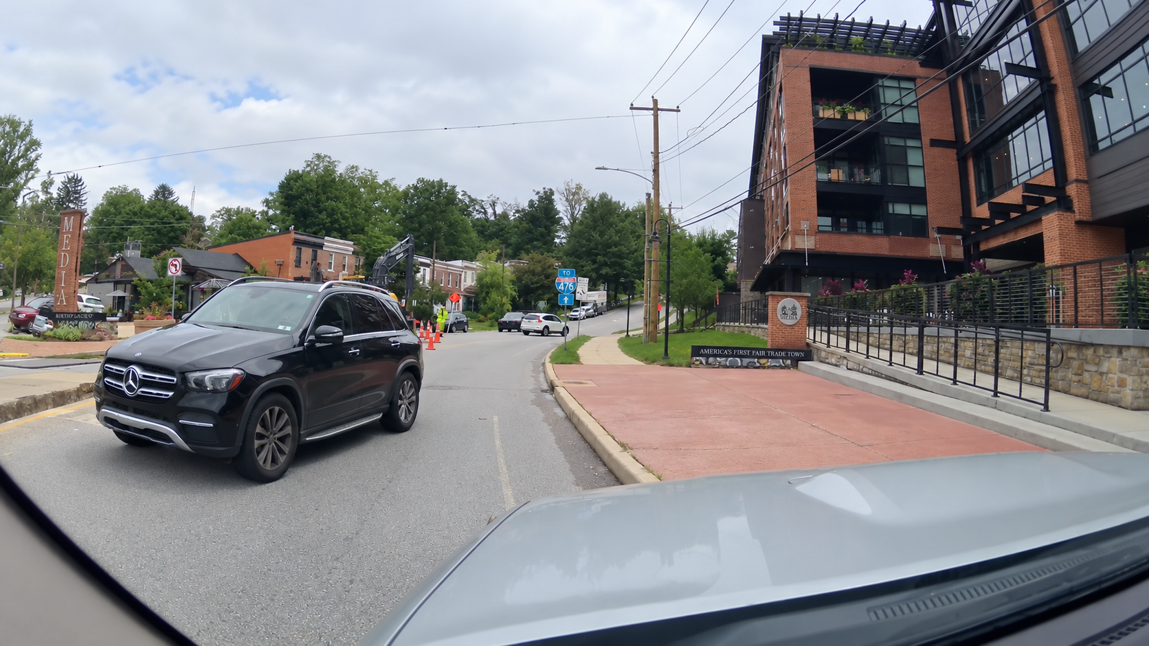A black SUV waits at an intersection near a modern brick building, with construction cones and workers visible on the road ahead. Trees and houses are in the background.