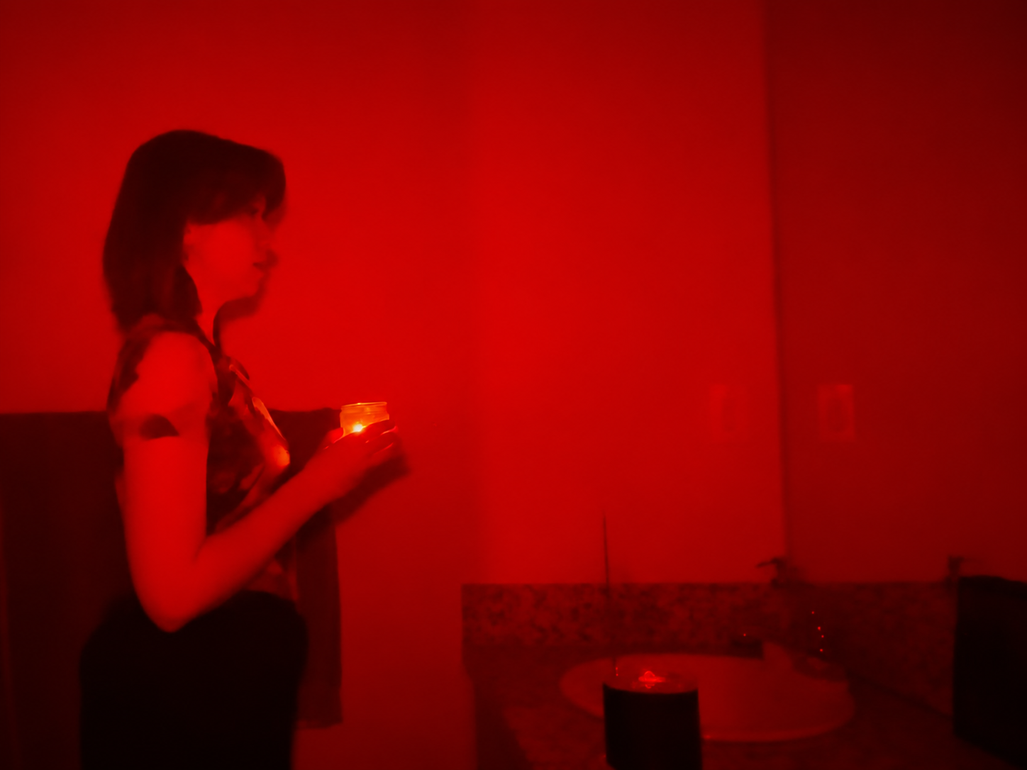A person holding a glowing candle stands by a bathroom sink, bathed in red light, with their reflection visible in the mirror.