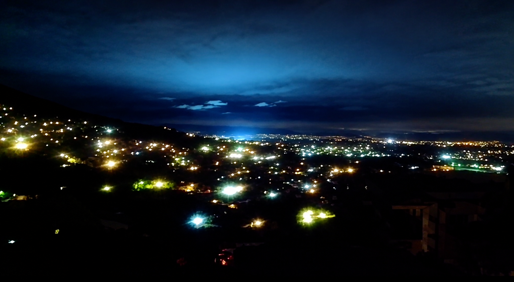 A cityscape at night with scattered lights glowing under a cloudy sky, hills in the foreground, and a blue glow illuminating the horizon.