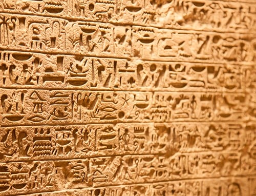 Close-up of an ancient stone wall covered in detailed Egyptian hieroglyphics, carved in horizontal rows and illuminated by warm light.