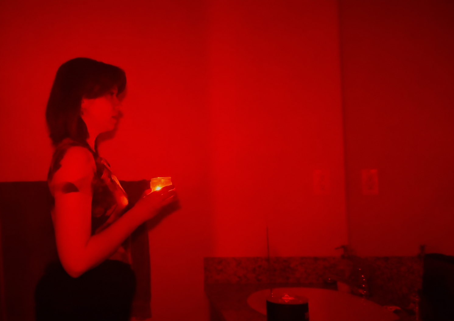 A person stands in a dim, red-lit bathroom holding a glowing candle near a sink and mirror. The atmosphere is eerie and mysterious.