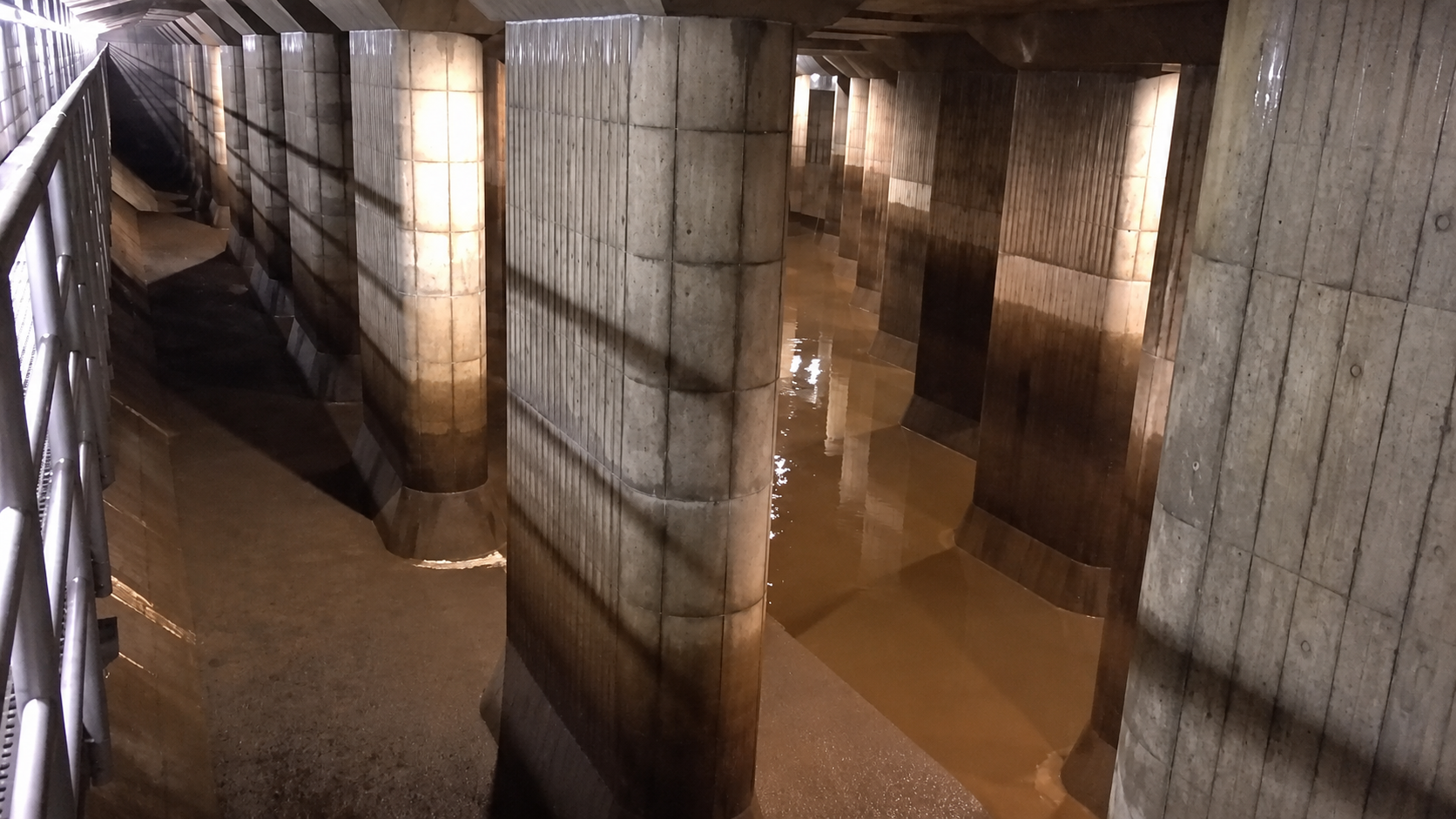 Concrete pillars support a bridge or overpass above shallow, muddy water. The scene is lit by artificial lights, casting shadows on the pillars. A metal railing runs along the left side.