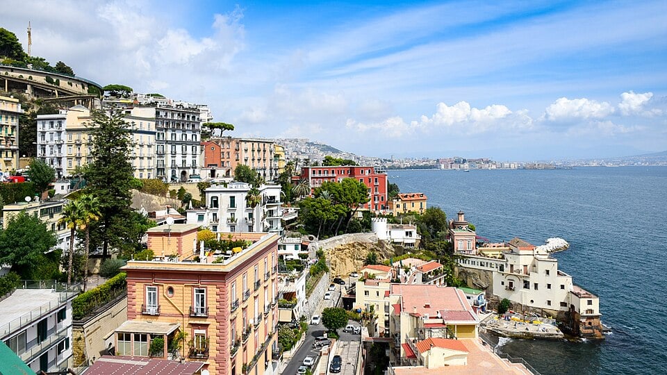 Colorful buildings on a hillside overlook the blue sea under a partly cloudy sky, with lush greenery and a coastal road winding along the shoreline.
