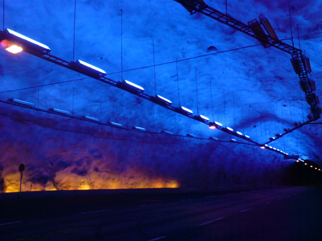 A tunnel illuminated with blue and purple lights. The ceiling appears textured, and the road is empty, creating a surreal, futuristic atmosphere. Yellow-orange lights line the lower part of the tunnel walls.