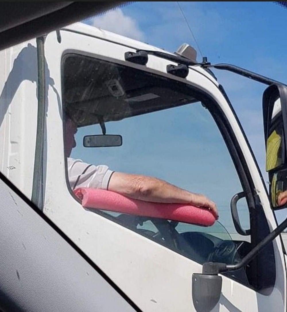 A driver rests their arm on a pink pool noodle used as an armrest inside a white truck. The image is taken from another vehicle, with part of its window and door visible in the foreground.