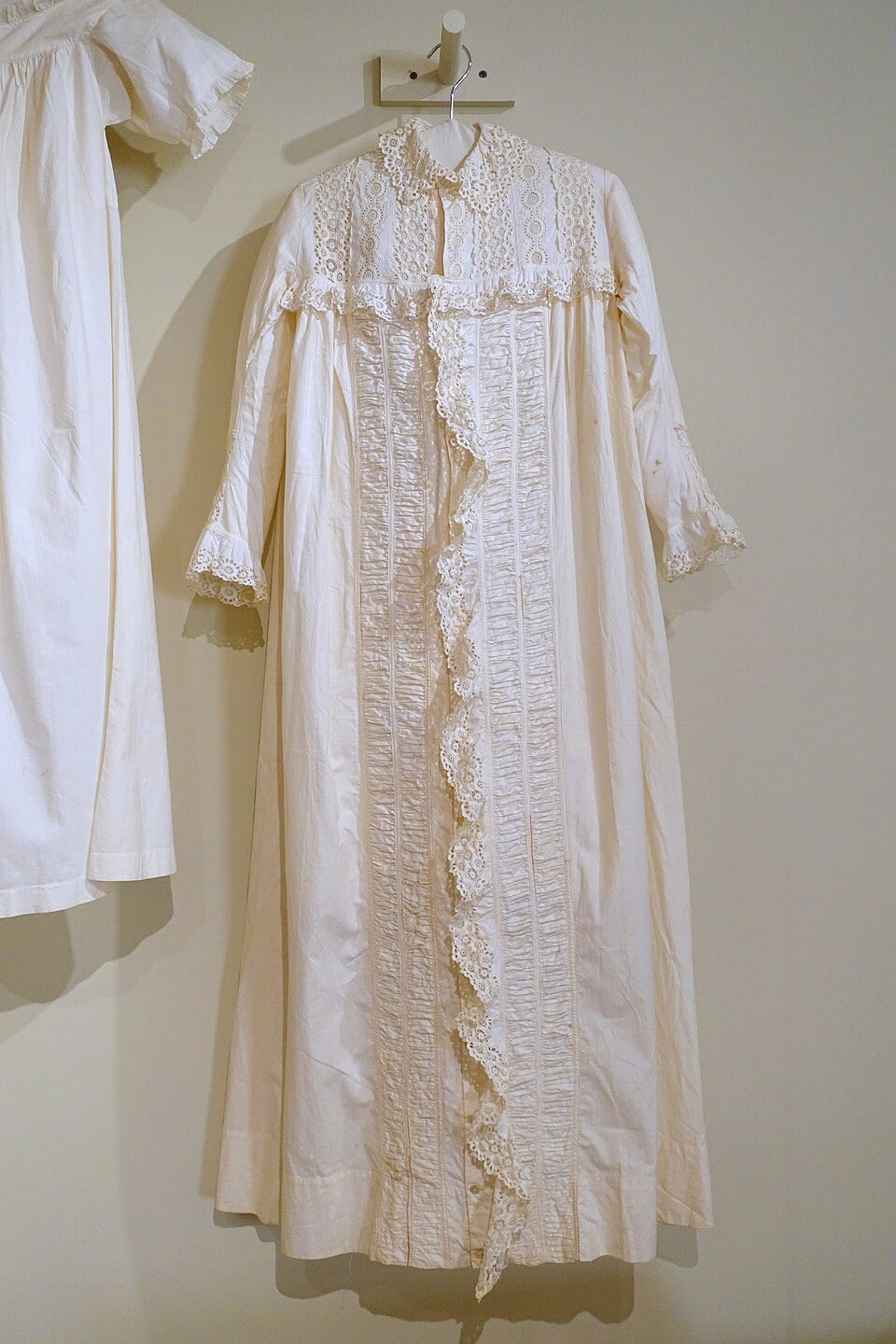 A long, vintage white lace nightgown with ruffled trim and decorative embroidery hangs on a wall hook, next to another white garment.