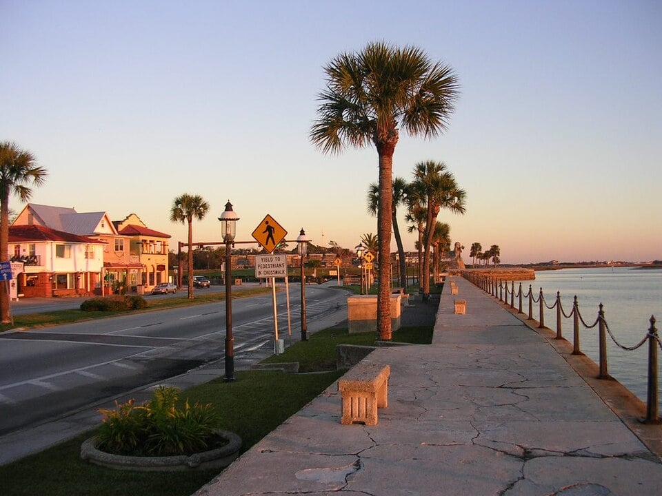 A waterfront street with palm trees, benches, street lamps, and pedestrian signs at sunset. Historic-looking buildings line the opposite side of the road.