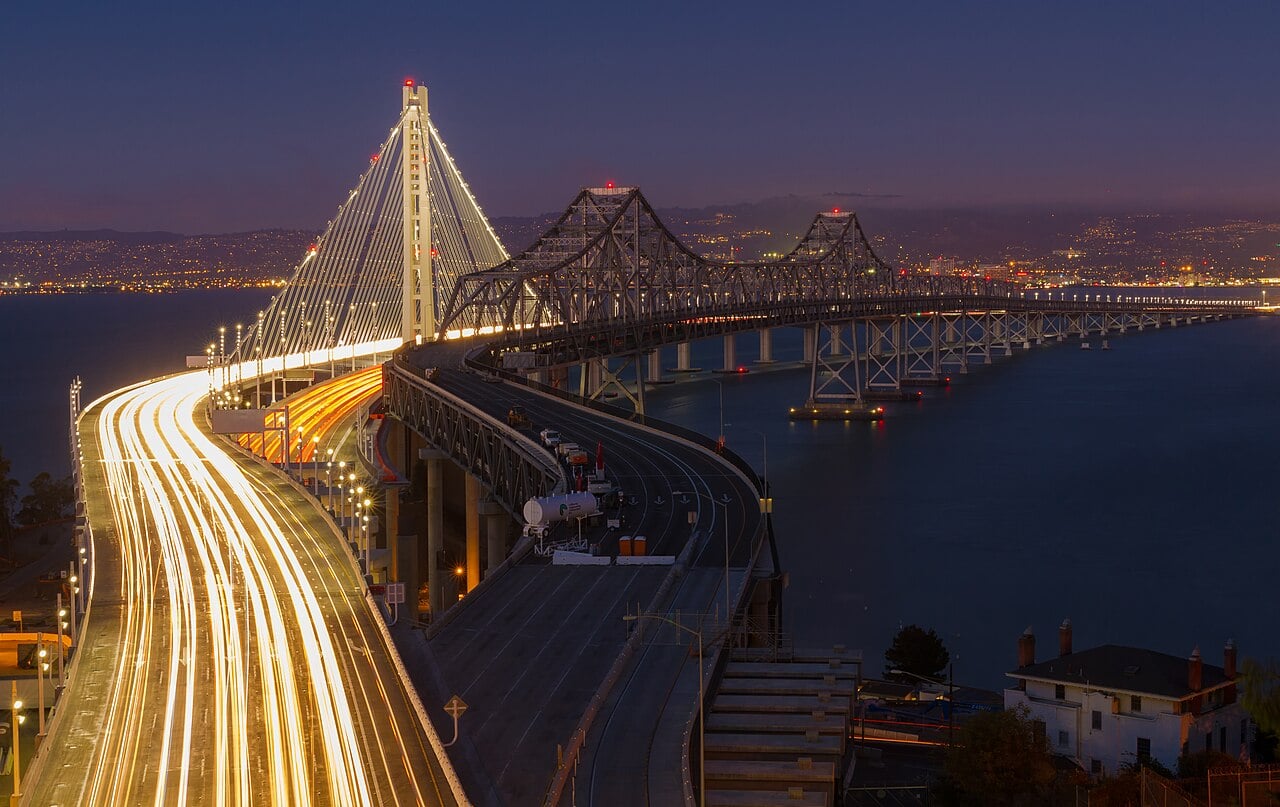 The Bay Bridge at night with light trails from cars, illuminated bridge spans, and city lights in the background over the water.
