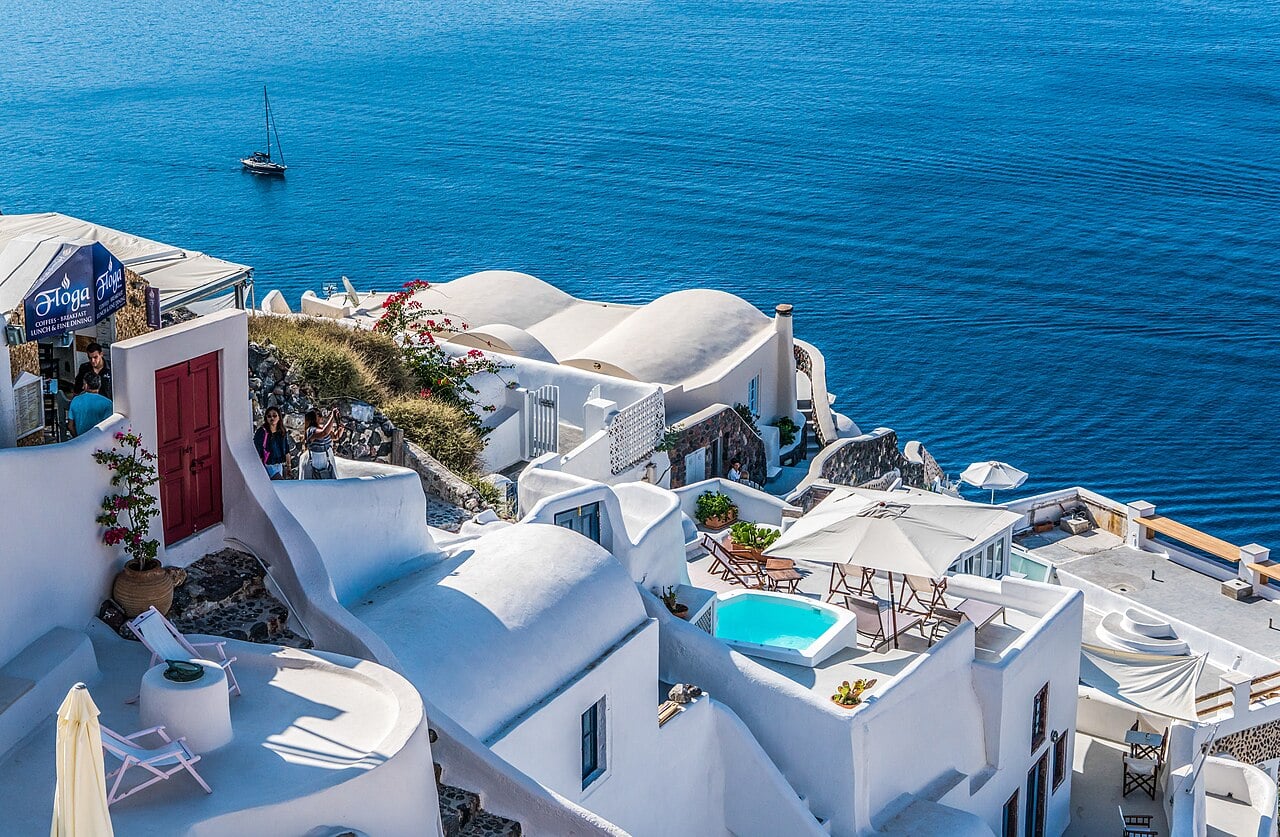 White buildings with blue domes and terraces overlook the calm, deep blue sea. A sailboat floats in the distance. There are pools, umbrellas, and flowers on the cliffside in bright sunlight.