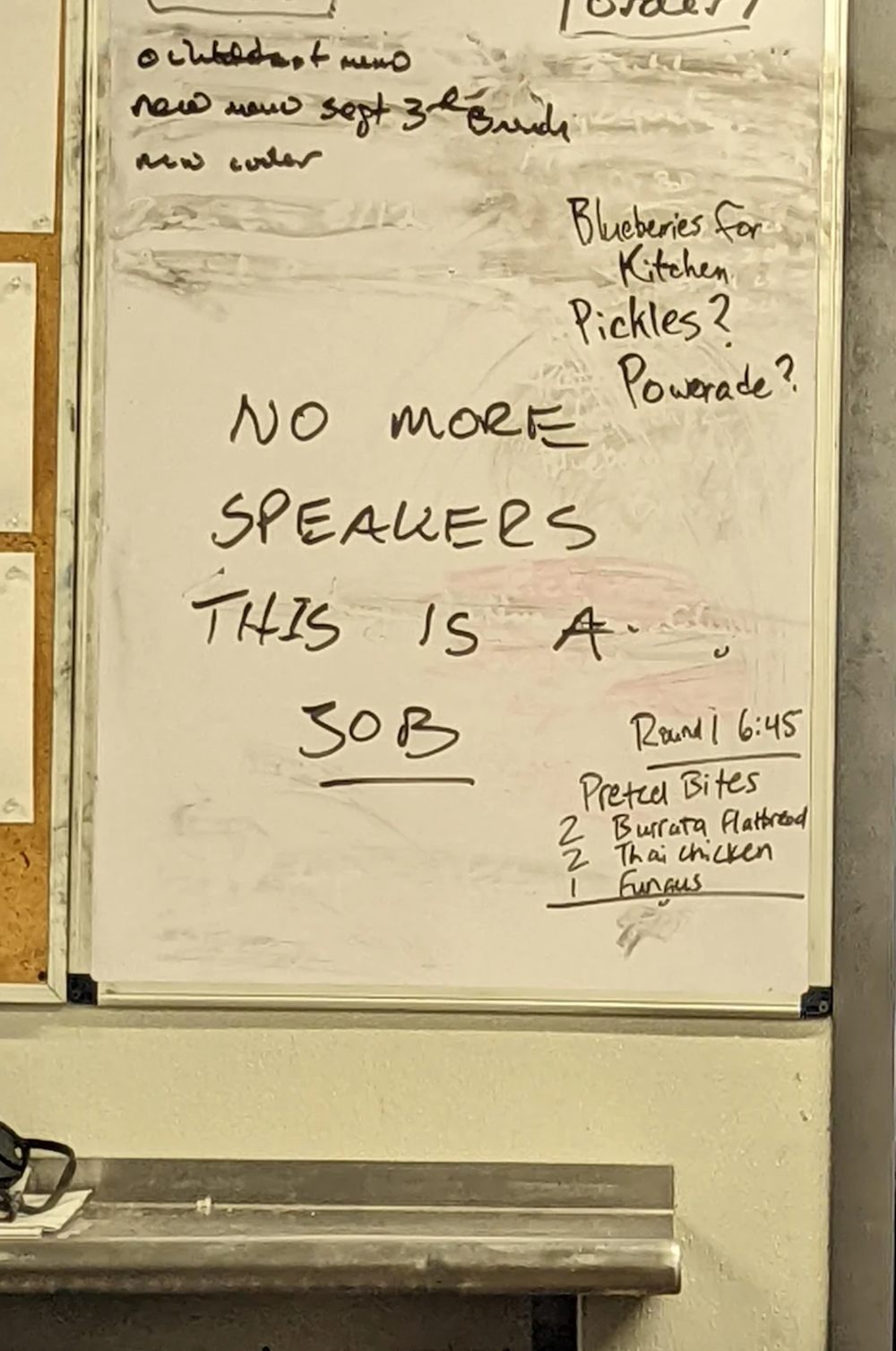 A whiteboard with handwritten notes, including “NO MORE SPEAKERS, THIS IS A JOB.” There are also grocery items like blueberries, Powerade, and a list with “Pretzel Bites, Burrata Flatbread, Thai Chicken Tacos.”
