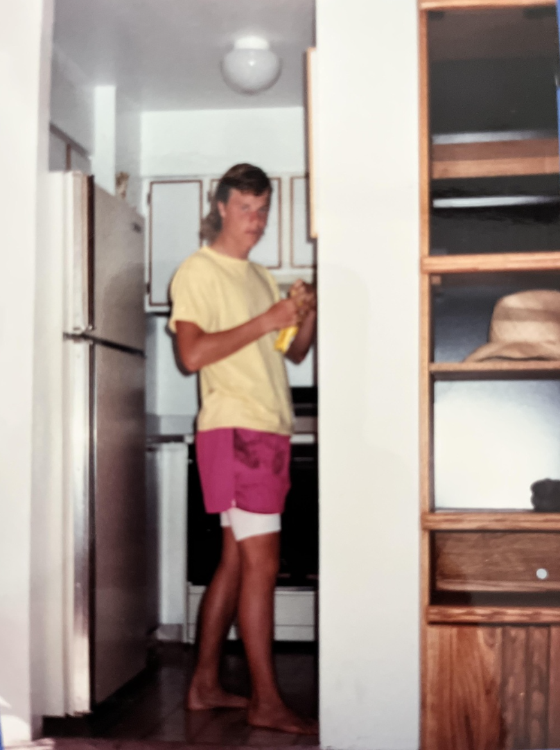 A person in a yellow shirt and pink shorts stands barefoot in a kitchen, holding a yellow item. Shelving and a refrigerator are visible.