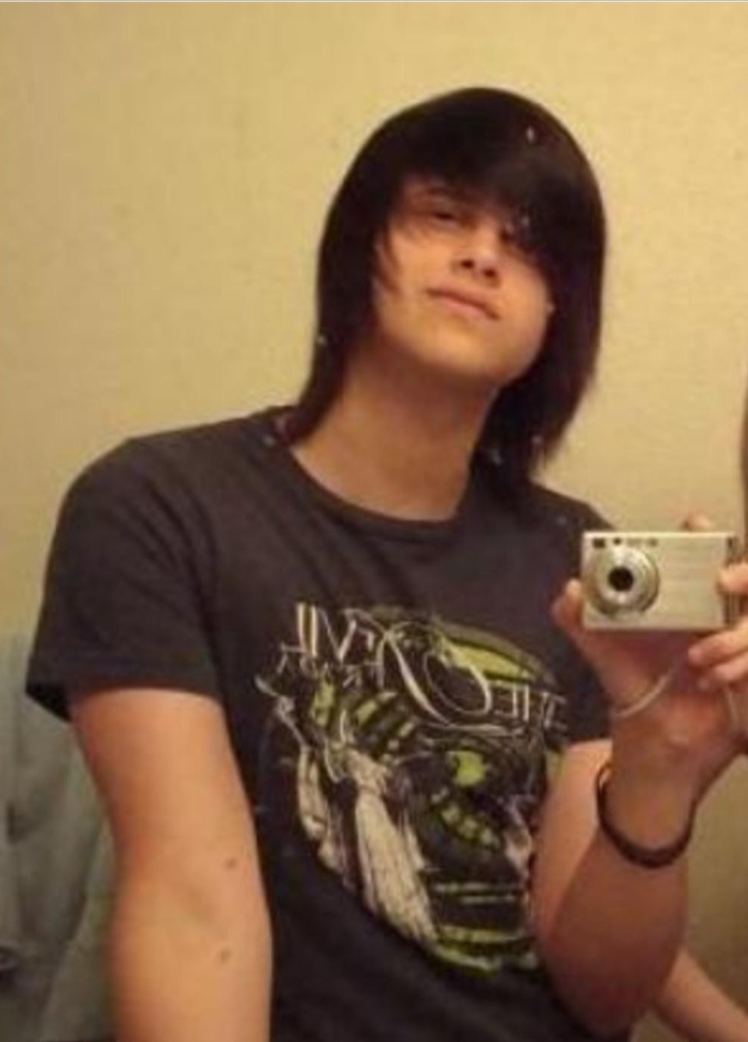 Young person with straight, dark hair covering one eye takes a mirror selfie with a digital camera, wearing a graphic t-shirt and standing in front of a plain wall.