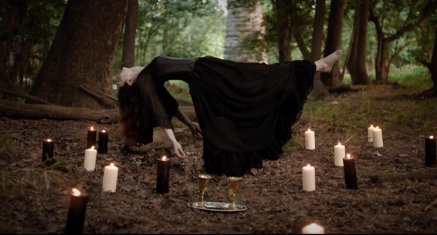 A woman in a black dress levitates horizontally above the forest floor, surrounded by lit white and black candles arranged in a circle. A tray with two glass goblets sits nearby.