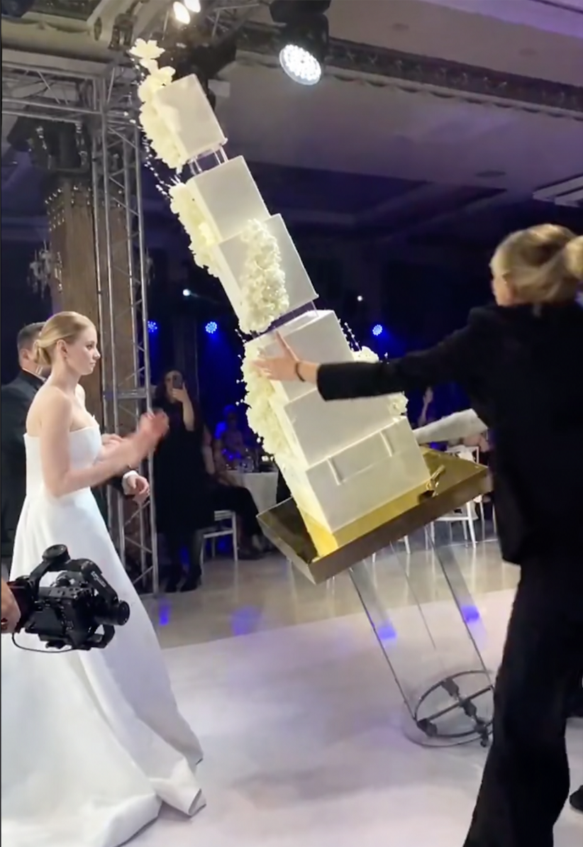 A large, tiered wedding cake is tipping over as a person tries to stop it from falling. A person in a white dress and a videographer stand nearby, capturing the dramatic moment.