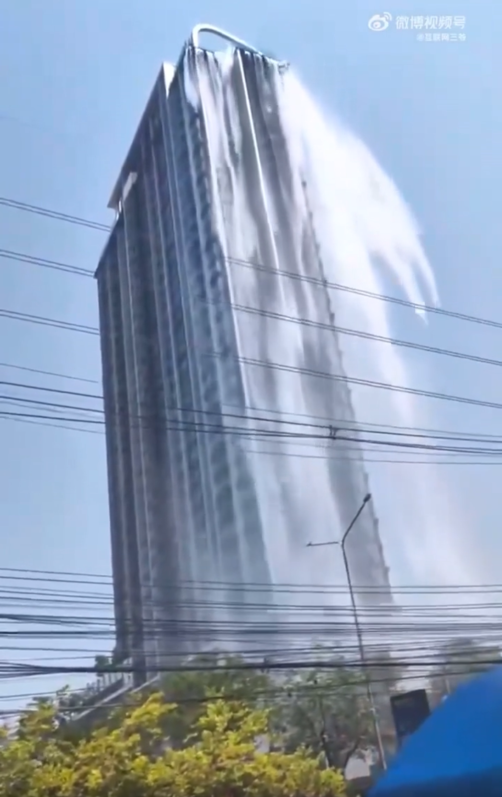 A tall building has water dramatically cascading from its roof, resembling a waterfall. Power lines and trees are visible in the foreground under a clear sky.