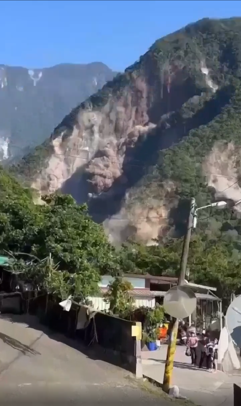 A landslide descends a mountainside near a village. People stand outside, watching as dust rises. The mountain is steep with patches of trees, and houses with satellite dishes are visible in the foreground.