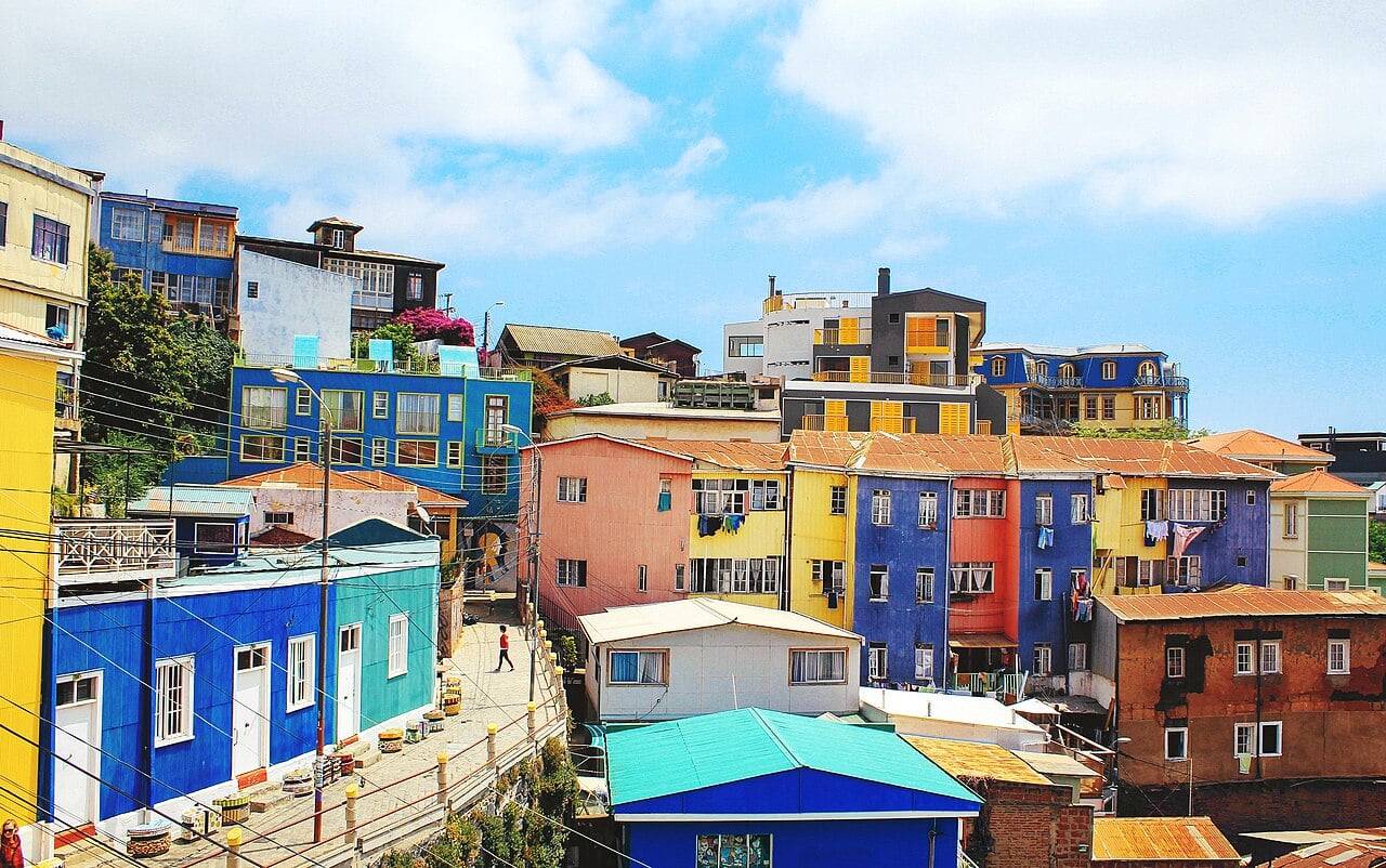 Colorful houses line a hillside under a partly cloudy sky. People walk along a narrow street, and laundry hangs outside. The buildings are painted bright blue, orange, yellow, pink, and gray, creating a vibrant neighborhood scene.