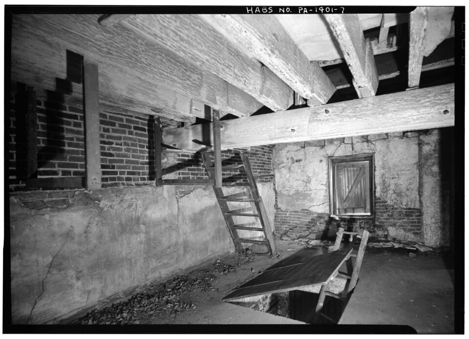 Black and white photo of a basement with wooden beams, brick and concrete walls, a wooden ladder, a small window, and a broken wooden table on the ground. The setting appears old and abandoned.
