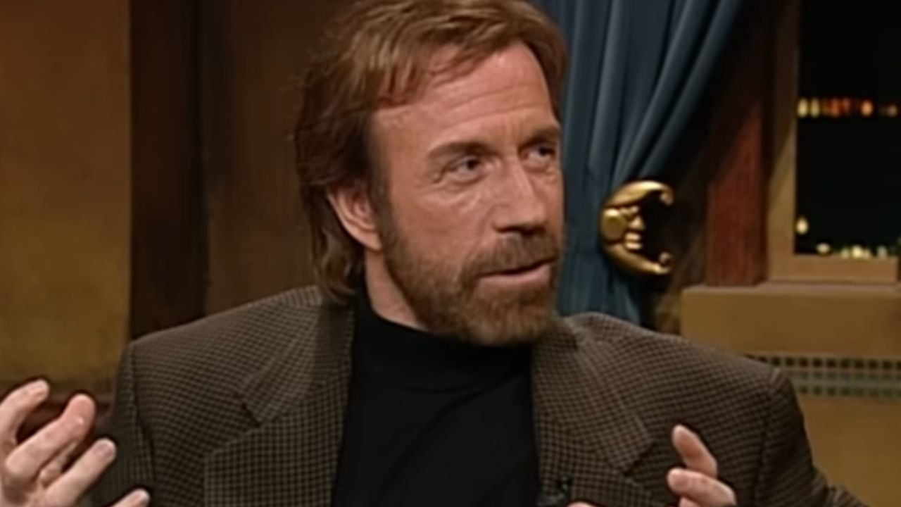 A man with light brown hair and a beard, wearing a dark turtleneck and checkered blazer, gestures with his hands while speaking indoors with a blue curtain in the background.