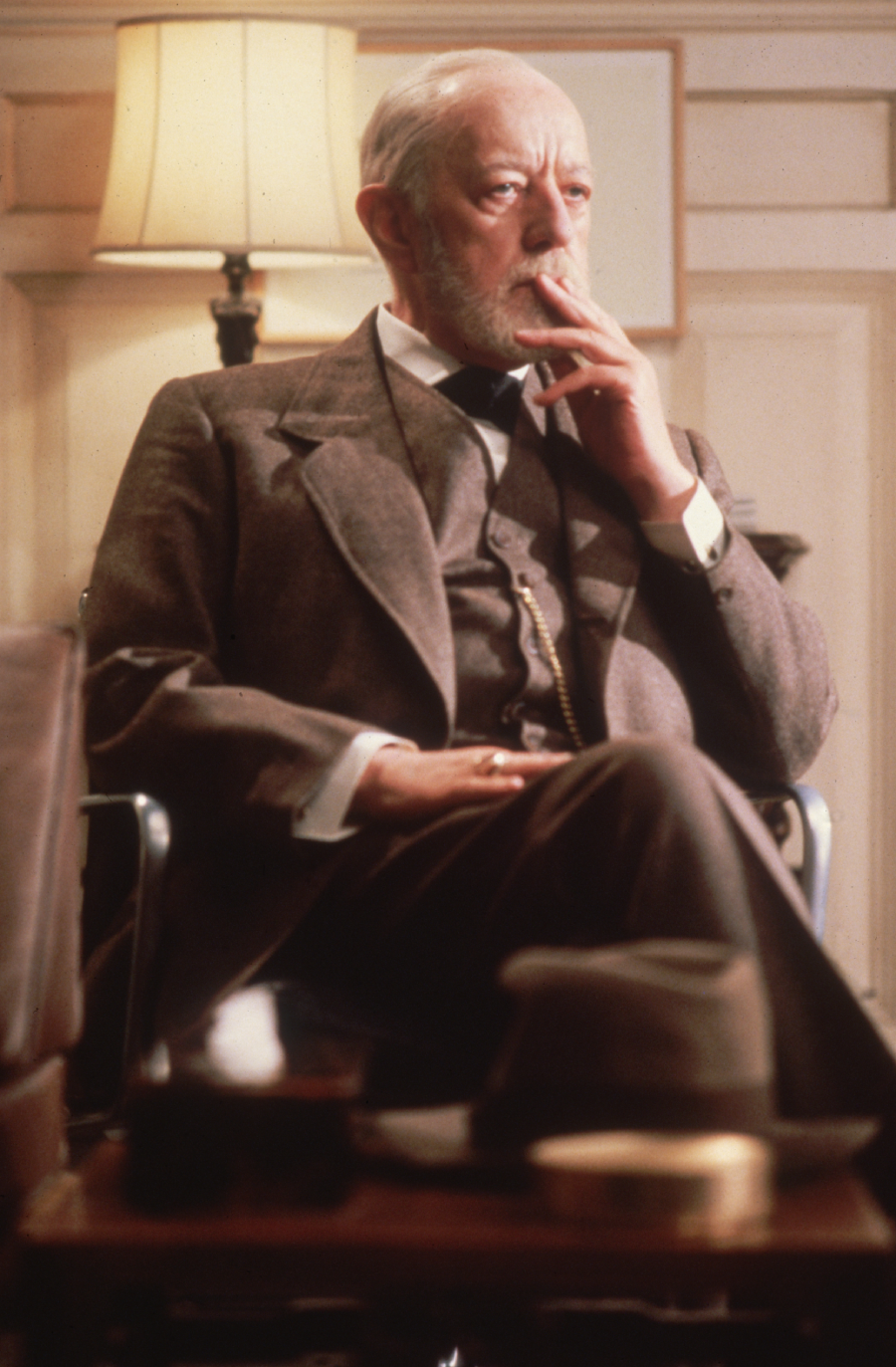 An elderly man in a brown suit sits in an armchair, thoughtfully resting his hand on his face. A lamp and paneled wall are visible behind him, and a table with a hat and objects is in the foreground.