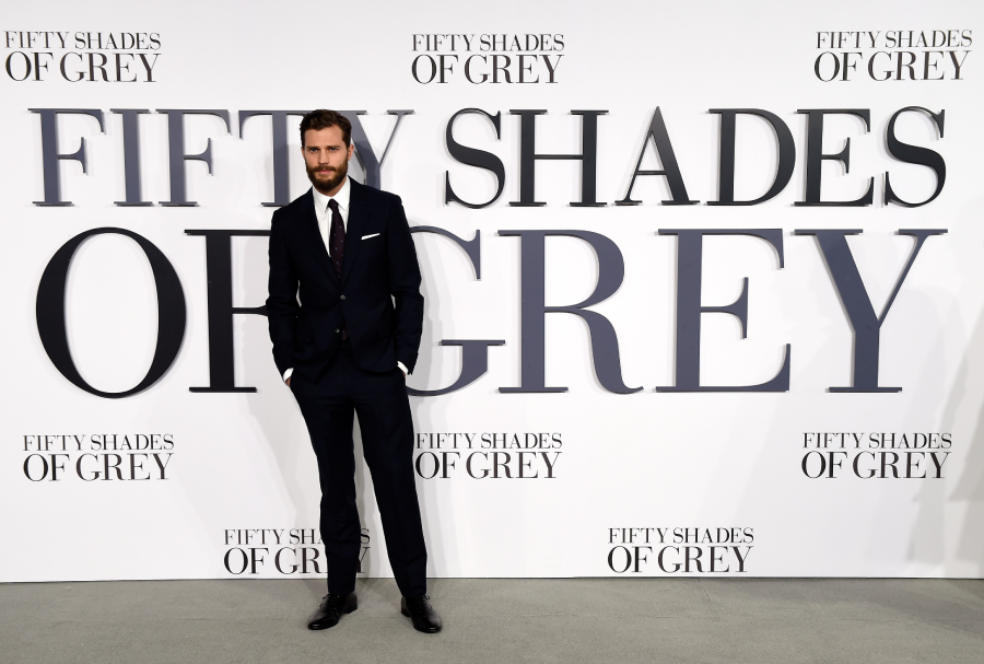 A man in a dark suit stands in front of a large “Fifty Shades of Grey” backdrop at a promotional event. The background features the movie title repeatedly in bold, black letters.
