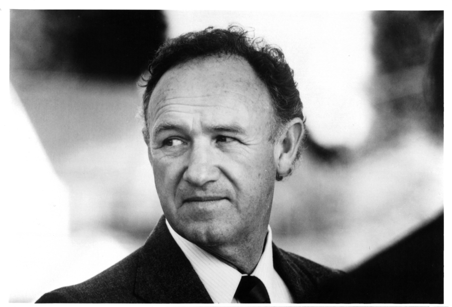 A man in a suit and tie looks to his left with a neutral expression. The photo is black and white, and the background is blurred.