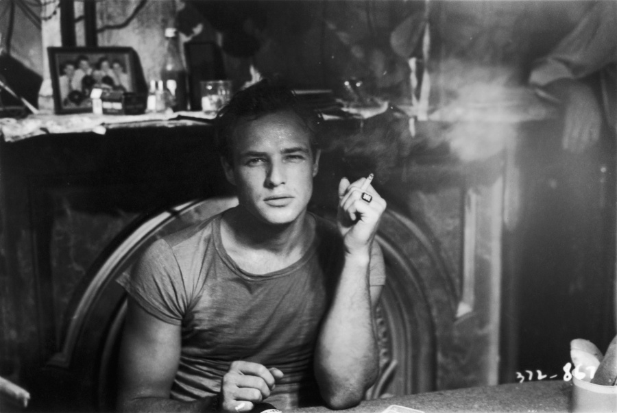 A man in a t-shirt sits at a table, holding a cigarette, with smoke rising. Behind him are framed photos, bottles, and clutter on a mantelpiece. The scene is in black and white.