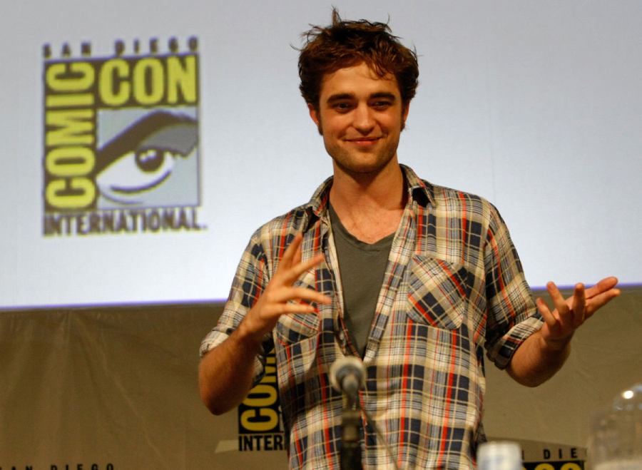 A person in a plaid shirt smiles and gestures while speaking at a San Diego Comic-Con International panel, with the Comic-Con logo visible in the background.