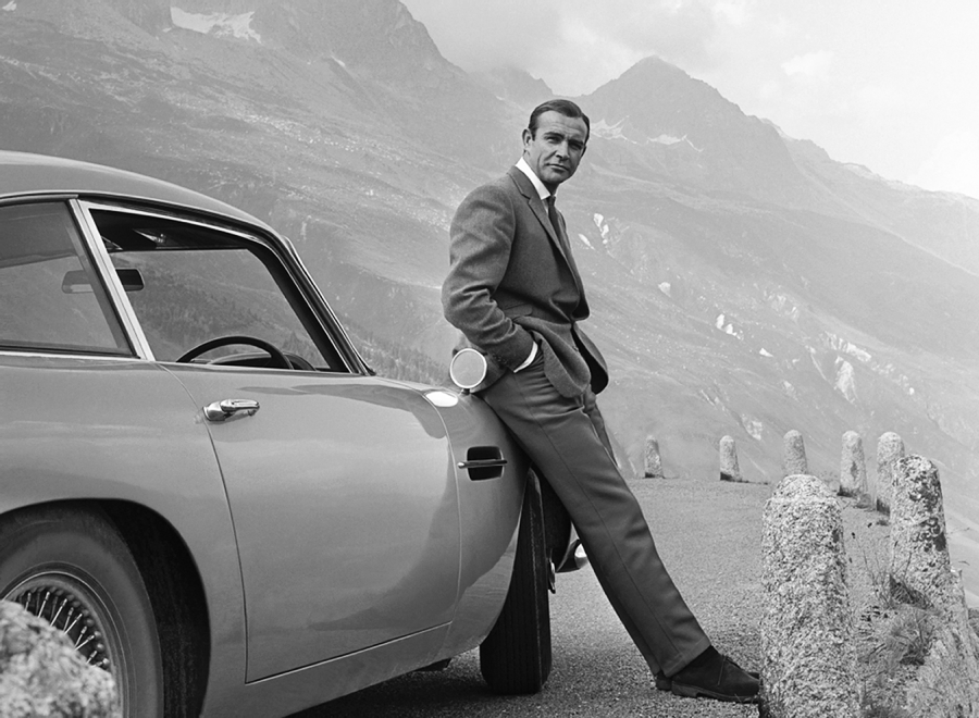 A man in a suit leans against a classic car parked on a mountain road, with mountains and clouds in the background. The image is black and white, creating a vintage, cinematic atmosphere.