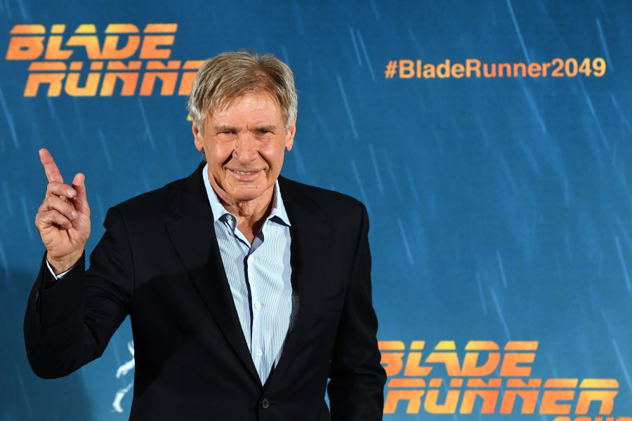 A man in a suit smiles and points upward on a red carpet in front of a “Blade Runner 2049” promotional backdrop.