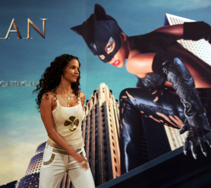 A woman in a white outfit walks past a large poster showing a costumed superhero crouching on a rooftop against a city backdrop.