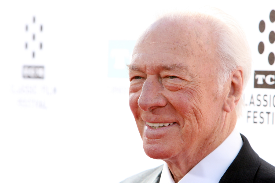 An older man with white hair smiles while wearing a light-colored suit and white shirt at an event with a white background and logos behind him.