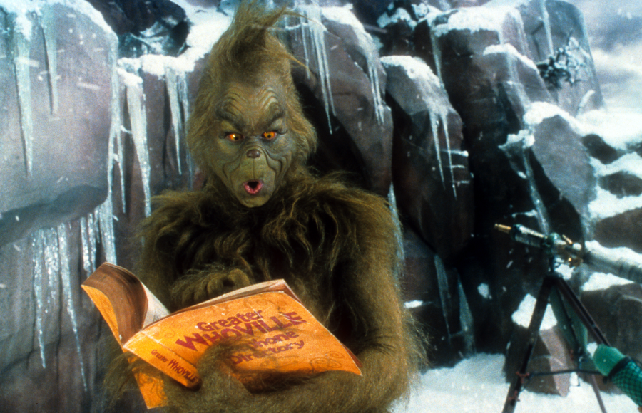 The Grinch, covered in green fur, stands in a snowy rocky landscape reading a book titled “Greater Whoville Directory” while a telescope is visible in the background.