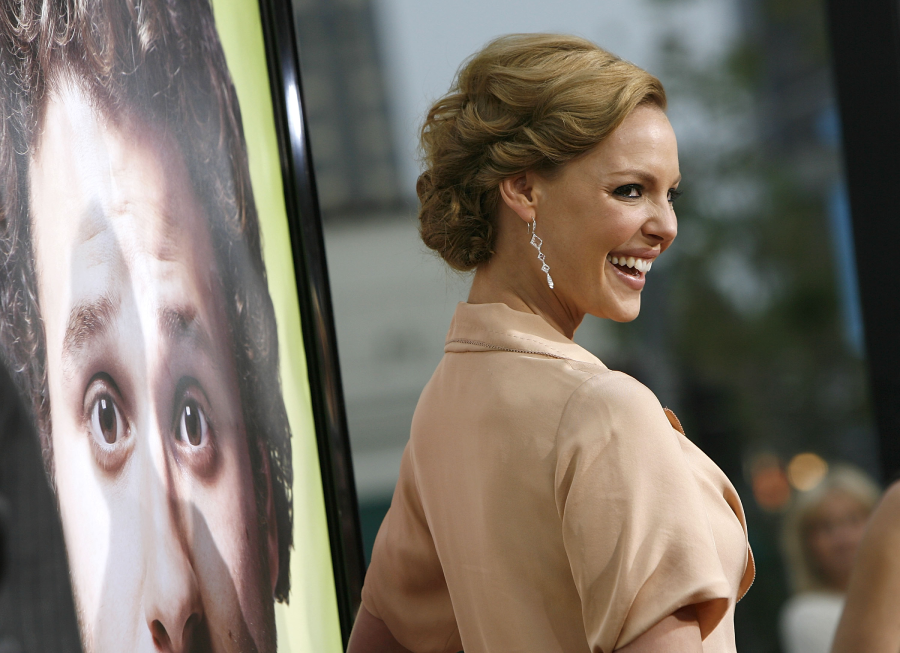 A woman in a light pink dress smiles over her shoulder at an event, standing next to a large poster showing a man’s surprised face.
