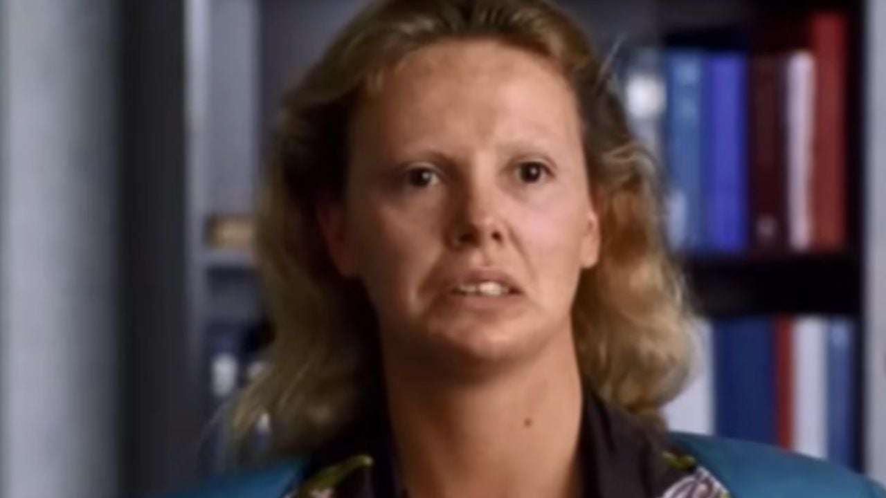 A woman with blonde hair and a serious expression faces the camera, with bookshelves blurred in the background.