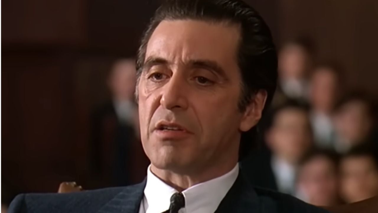 A man in a dark suit sits indoors, looking serious and focused. Several blurred people are visible in the background, suggesting a formal or courtroom setting.