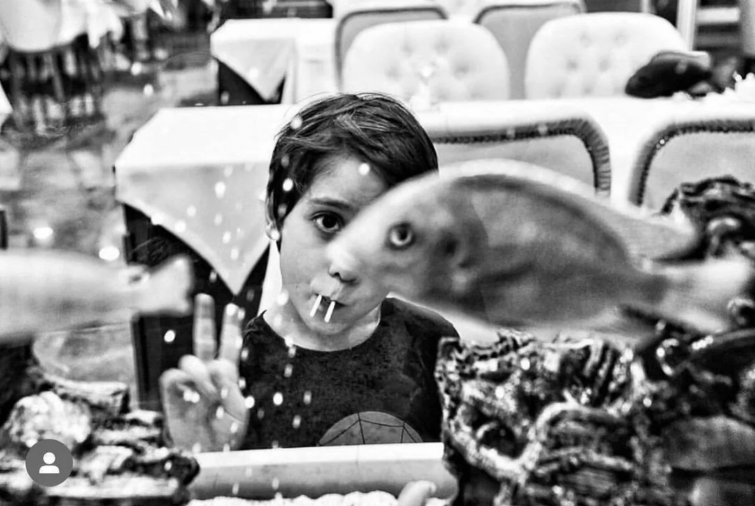 A child with candy sticks in their mouth looks through a fish tank, holding up a peace sign. Blurred fish swim in front. The image is in black and white.