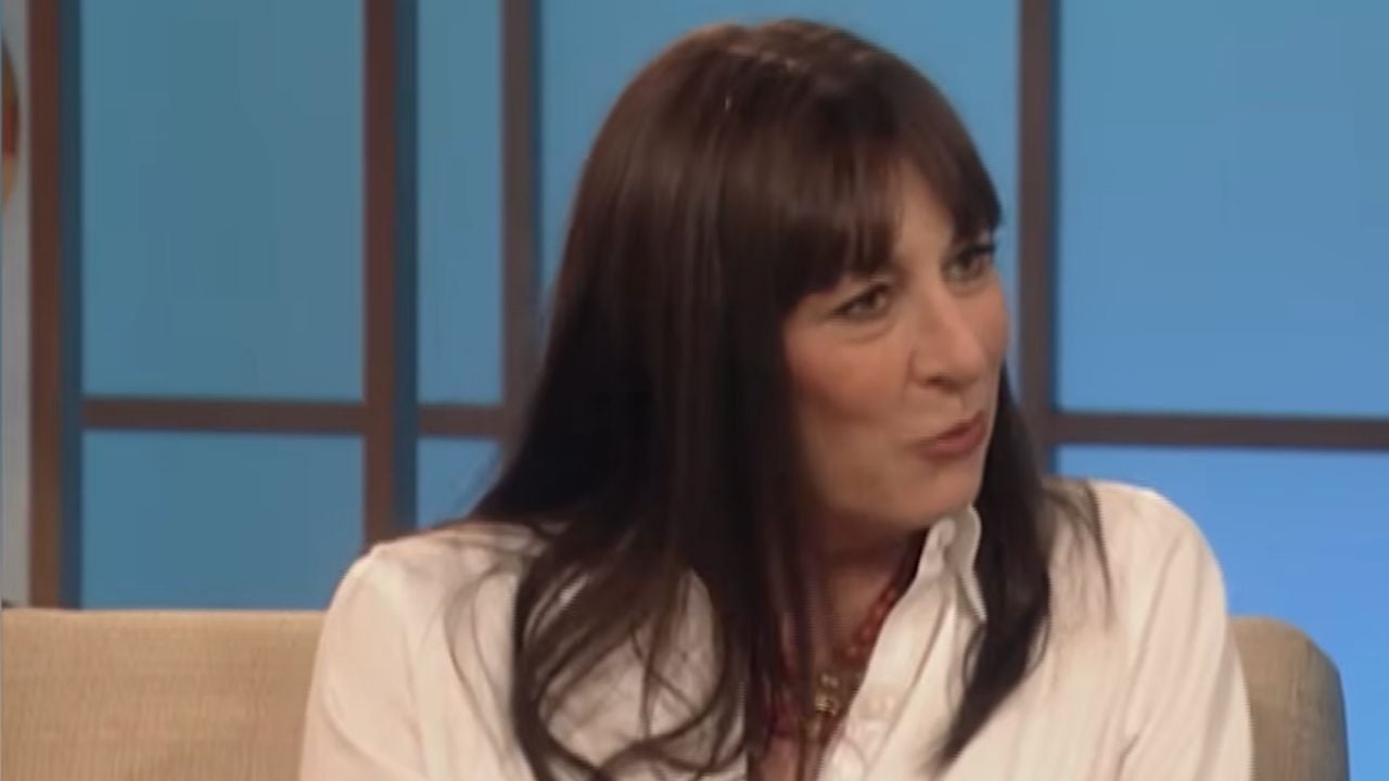 A woman with straight brown hair and bangs, wearing a white shirt, sits on a beige couch and talks. The background features blue panels.