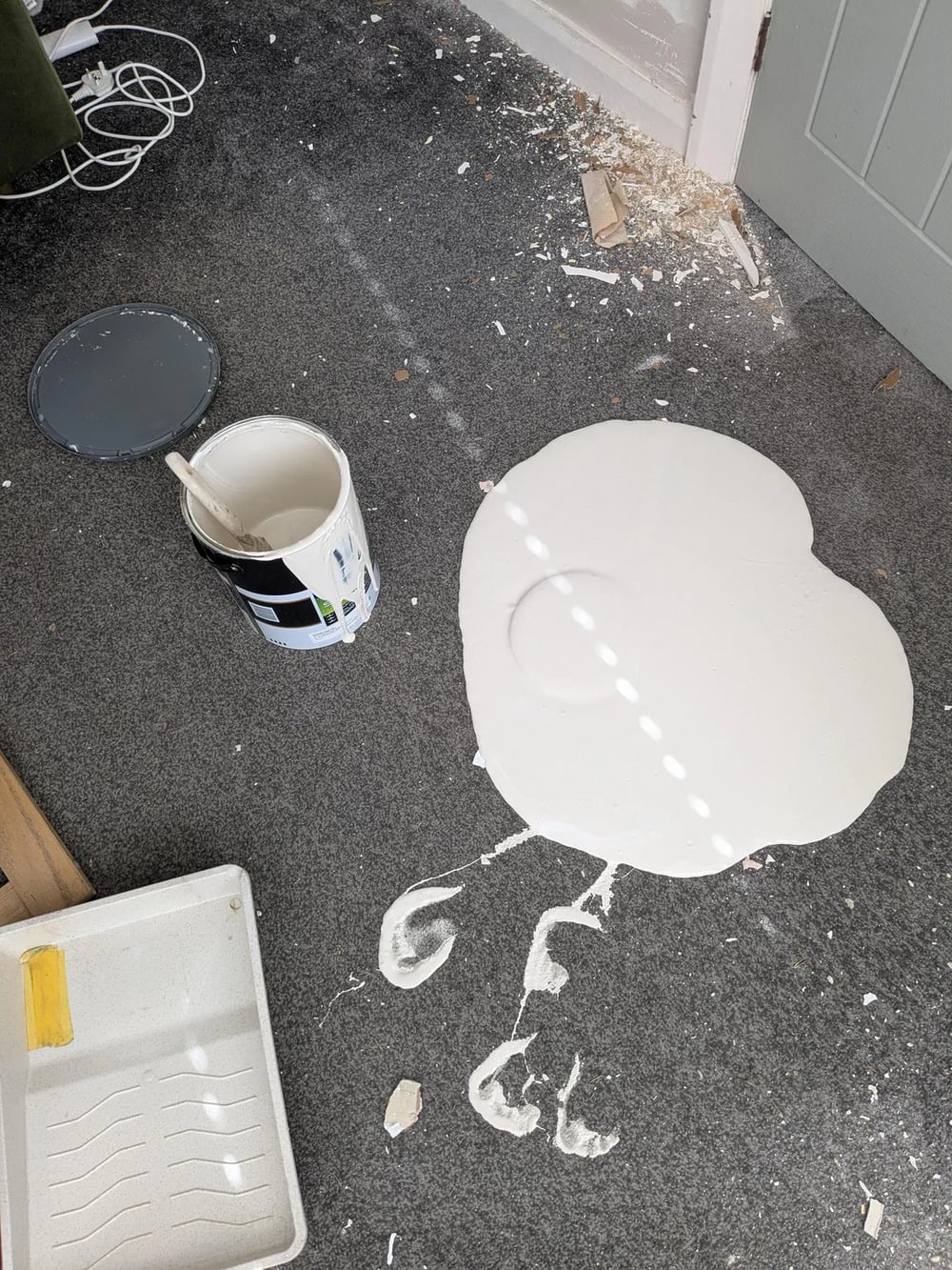 A large spill of white paint on a gray carpet, with an open, tipped-over paint can nearby and paint splatters around. A paint tray and scattered debris are also visible on the floor.