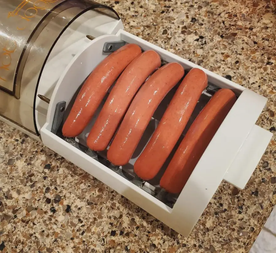 Five hot dogs are placed inside a white kitchen appliance on a speckled countertop. The appliance appears to be designed to hold and possibly cook or slice the hot dogs.
