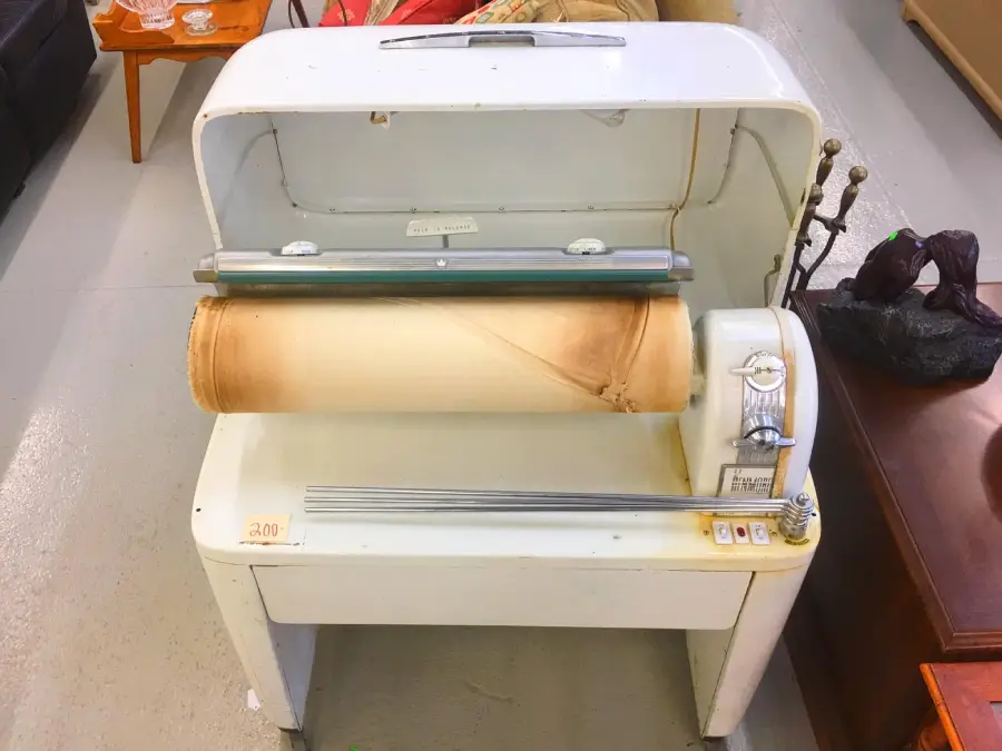 An old-fashioned electric mangle ironer with a large roller and metal cover, positioned on a white floor. The roller has a noticeable brown burn mark. Furniture and decor items are visible in the background.