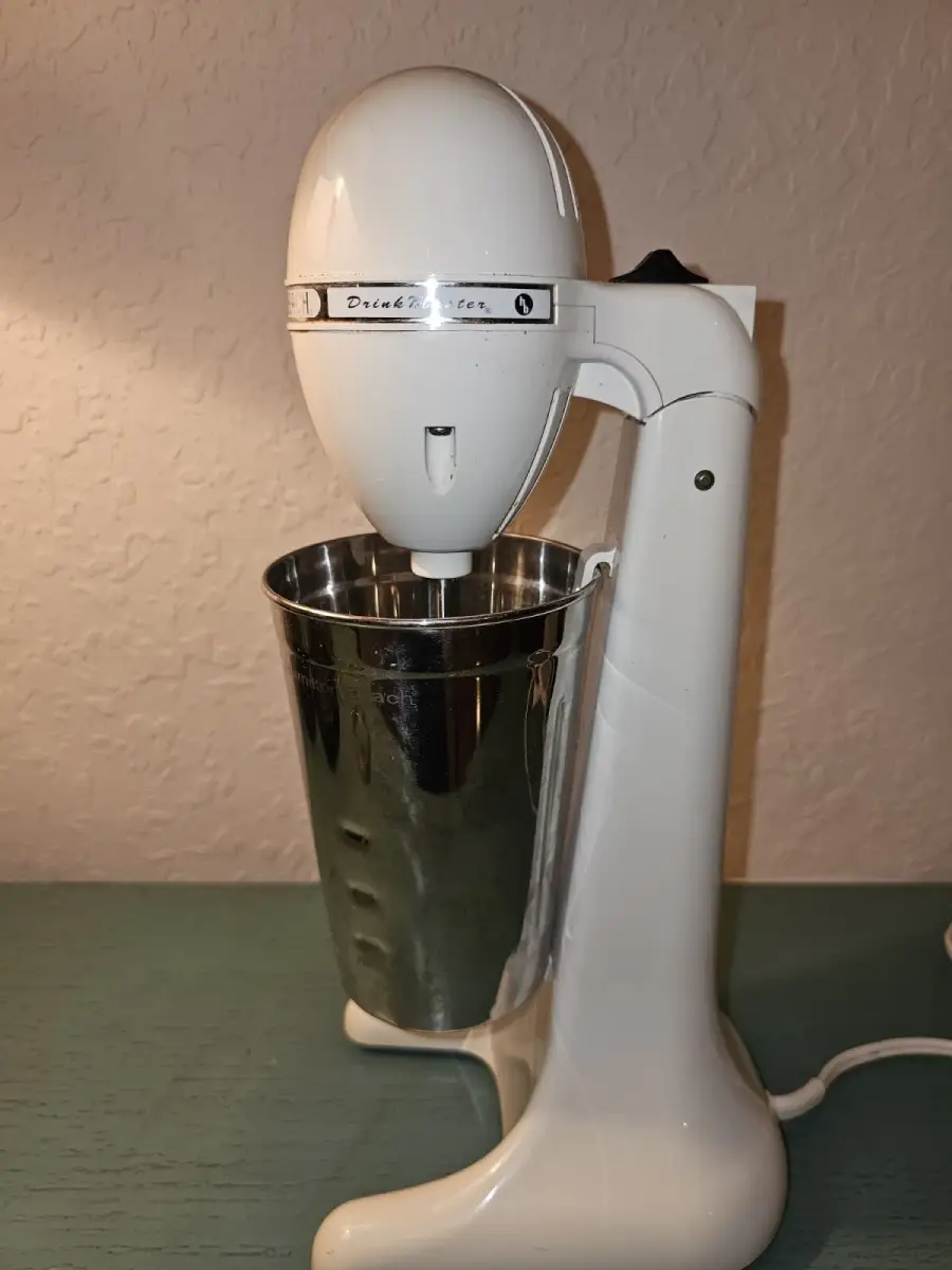 A vintage white milkshake mixer with a metal cup sits on a green surface against a beige textured wall.