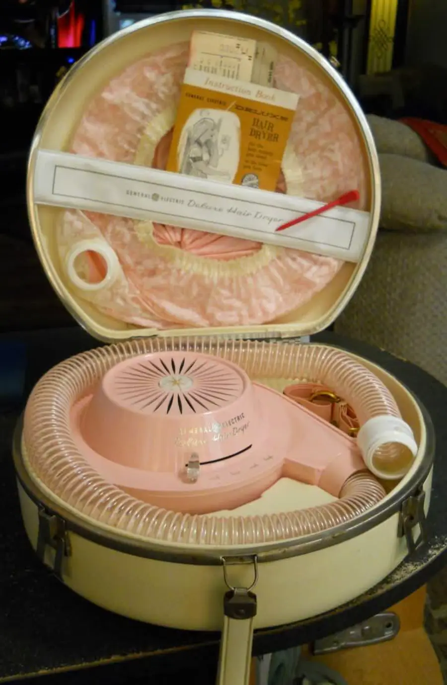 A vintage pink hair dryer with a hose, bonnet, and manual, all packed inside a round cream-colored carrying case.