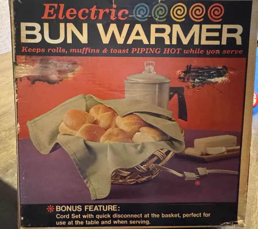Vintage packaging for an electric bun warmer, showing a basket with rolls covered by a cloth, a power cord, a coffee pot, and text promoting its features.