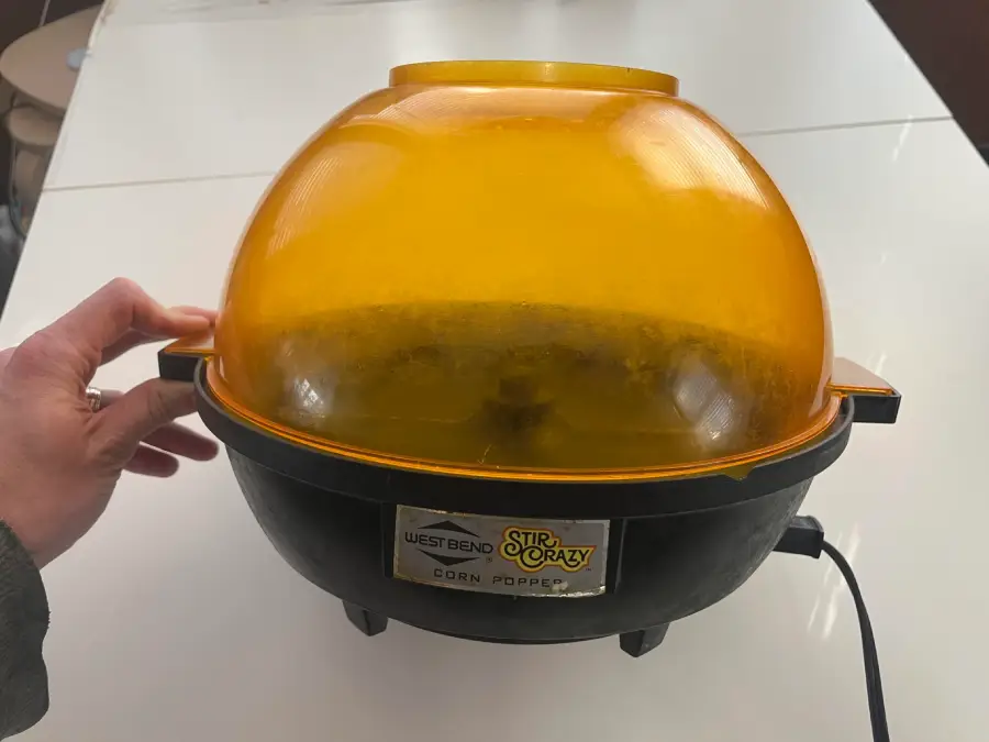 A hand holds a vintage West Bend Stir Crazy corn popper with a yellow dome lid, set on a white table.
