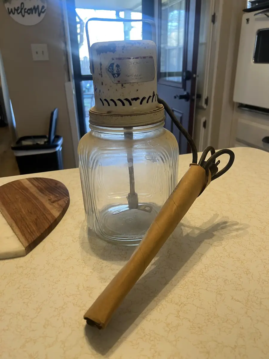 A vintage hand-crank glass butter churn with a metal lid and paddle mechanism sits on a kitchen counter. A rolled-up brown paper stick lies in front of it.