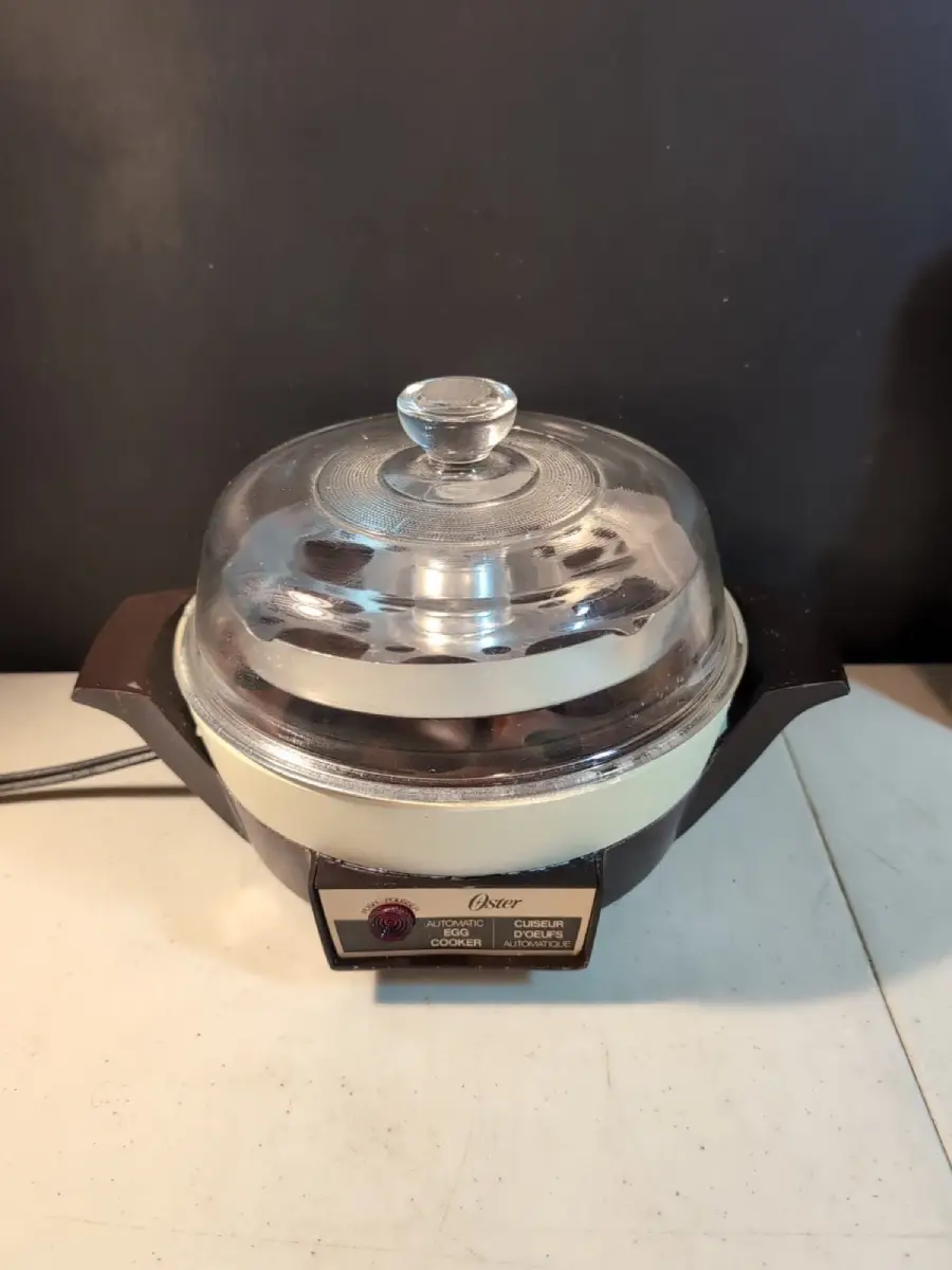 A vintage electric skillet with a clear glass lid, black handles, and a temperature control dial sits on a white surface against a dark background.