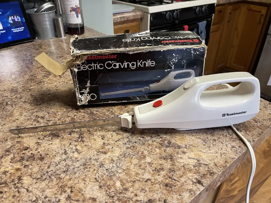 A white Toastmaster electric carving knife with serrated blades is on a kitchen counter, next to its original black box. The kitchen features brown cabinets and appliances in the background.