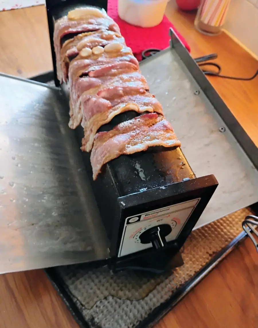 Raw bacon strips are draped over a black and silver bacon-cooking appliance on a countertop, with a dial on the front and kitchen items in the background.